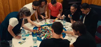 Leadership team collaborating during a strategy workshop, with LEGO models and discussion groups visible. Warm, authentic office environment reflecting teamwork, innovation and company culture.