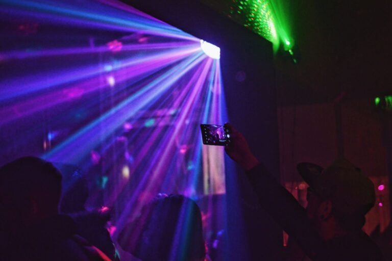 Neon lights illuminate parts of a dark room, and a man holds up his phone to capture the lighting.
