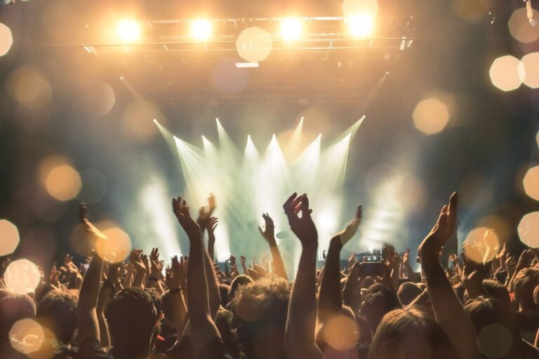 Bright arena and stage lights with a large crowd of people facing the stage, many with their hands in the air.