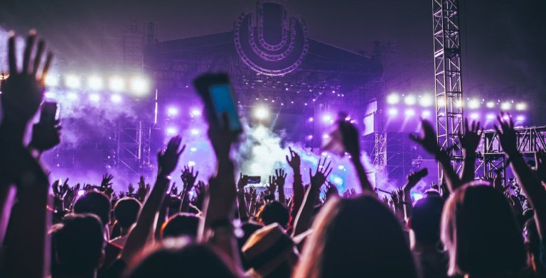 A huge crowd of people with their hands up, dancing and enjoying the music in front of a festival stage covered in purple lights.