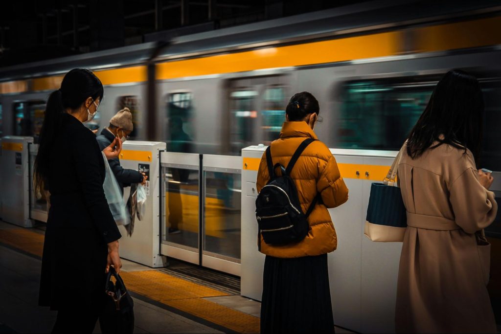 A diverse group of people stands at a train station, patiently waiting for their train to arrive.