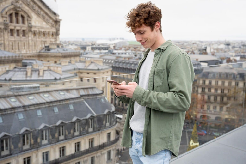 Young male with curly hair on a rooftop in a northern European city smiling while texting.