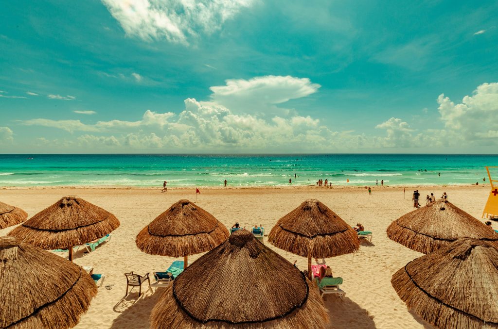 A view of the beach, including straw umbrellas, tourists lounging in the sun, and people playing in the ocean under the bright blue sky. 