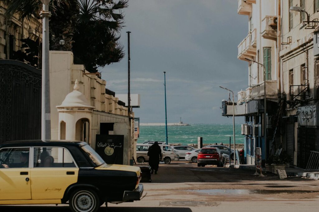 A black and yellow taxi drives down a street in Alexandria, Egypt, near the coastline, with historic buildings and the sea visible in the distance.