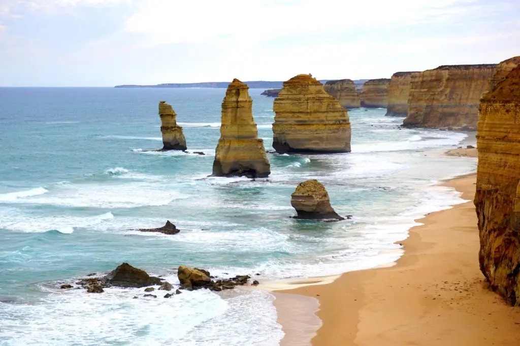 A view of the Twelve Apostles, limestone rock formations off the coast of the Great Ocean Road in Victoria, Australia, with waves crashing along the sandy beach.