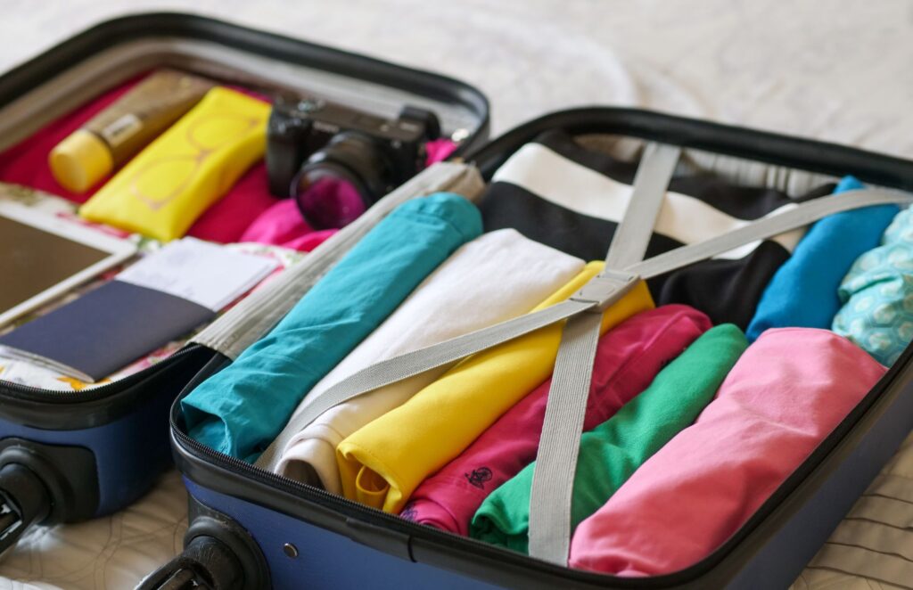 An open carry-on suitcase packed with tightly rolled, colorful clothing items, travel documents, a DSLR camera, sunscreen, and a pouch containing sunglasses.