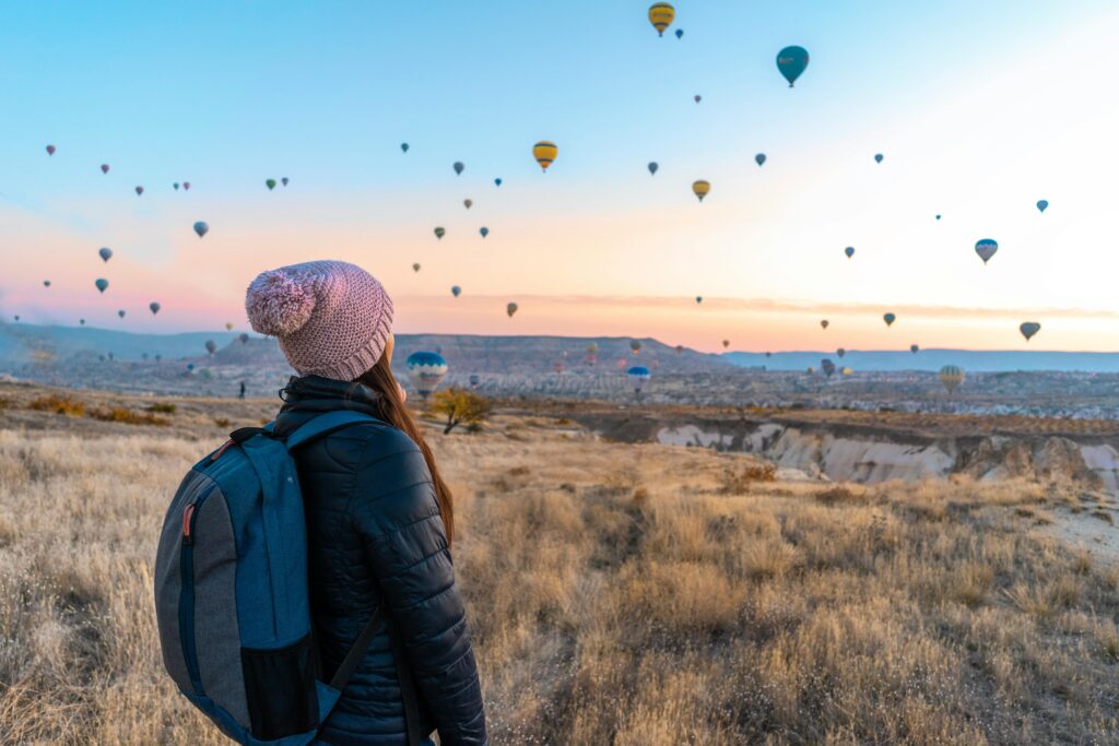 A photo of a female tourist hiking on the hills of Cappadocia, looking at the sky filled with colorful hot air balloons.