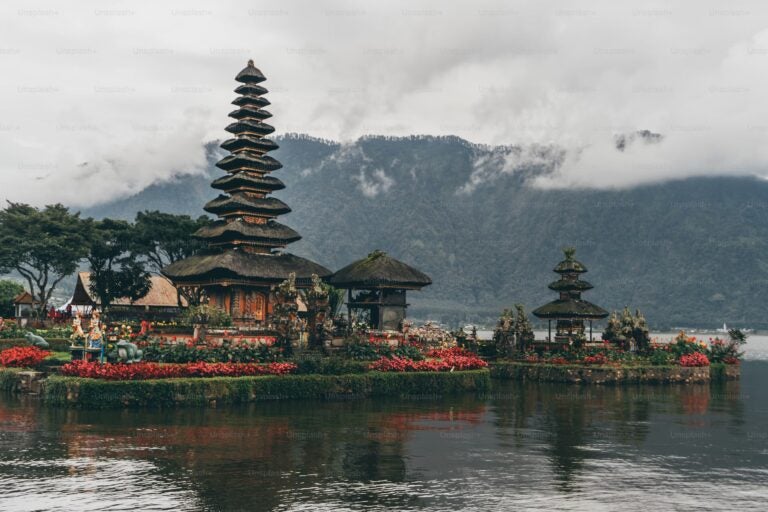Balinese Hindu temple with stacked roofs reaching towards the sky, surrounded by vibrant red flowers on the banks of a tranquil lake. Foggy clouds wrap around the distant hills covered in dense, dark green forests.