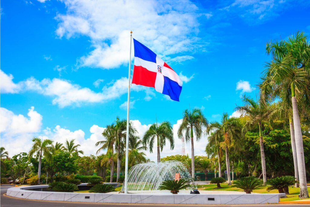 Image of Dominican Republic flag amid palm trees and blue and cloud-filled sky, Punta Cana.