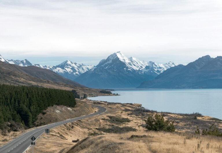 Scenic view of snow-covered mountain peaks in New Zealand with a winding road descending downhill, surrounded by hills, forests, and a lake.