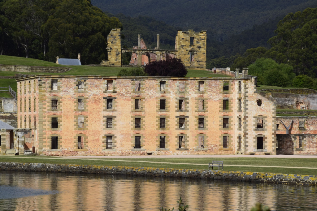 The historic ruins of Port Arthur by the shore against a dense dark green forest in the background.
