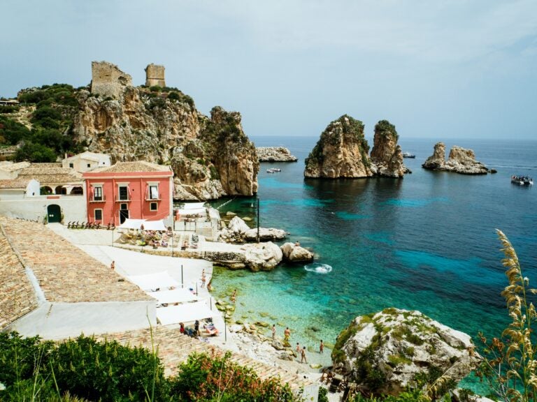 A beach in Sicily, Italy, with colorful buildings in the distance and rocky cliffs covered in lush greenery in the backdrop.

