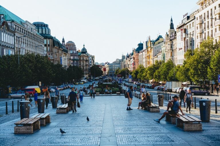 Travelers and locals relaxing in a square along a boulevard in Prague, with a mix of old and modern buildings forming a contrasting skyline against a clear sky