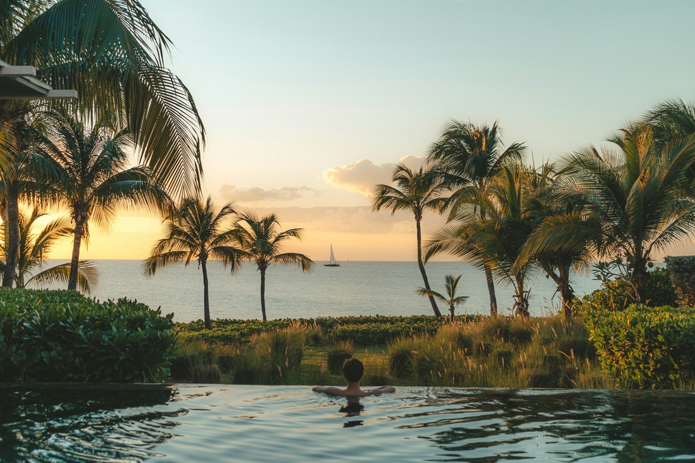 A man gazes over an infinity pool at a yellow sunset in Anguilla surrounded by palm trees 