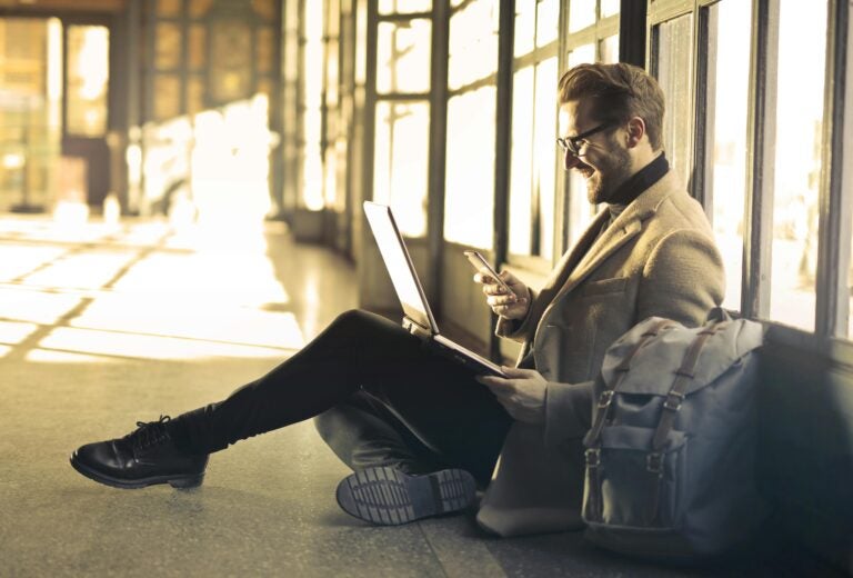 A man sitting in the airport corridor using his laptop and mobile device.