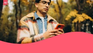 Young traveler holding a mobile device with an orange protective case against a blurry background of a forest in the fall.
