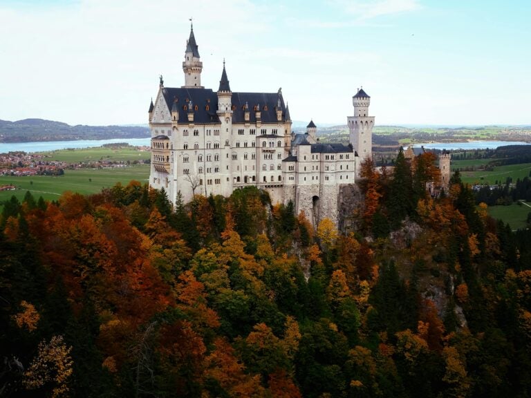 Neuschwanstein Castle in Germany, towering over a forest with red, ochre, and green foliage.