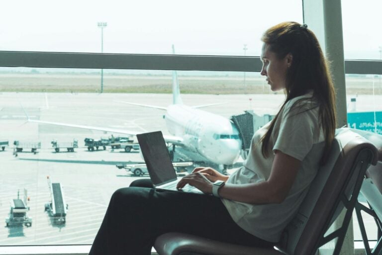 A woman sitting by the window at the airport, working on her laptop with an airplane in the background.