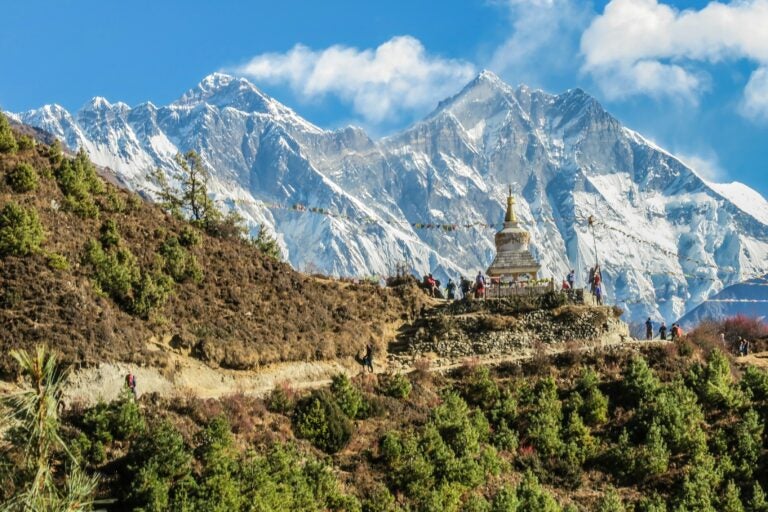 A narrow mountain trail in Nepal on a sunny day, with snowy mountain cliffs in the background.
