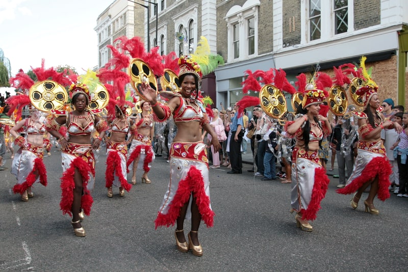 Dancers in elaborate costumes with red feathers at the Notting Hill Carnival Parade, with curious onlookers watching.
