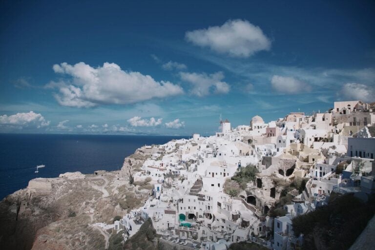 A view of Santorini, Greece, featuring its iconic chalk-white buildings built into the rocky hills, overlooking the deep blue sea on a bright, sunny day.