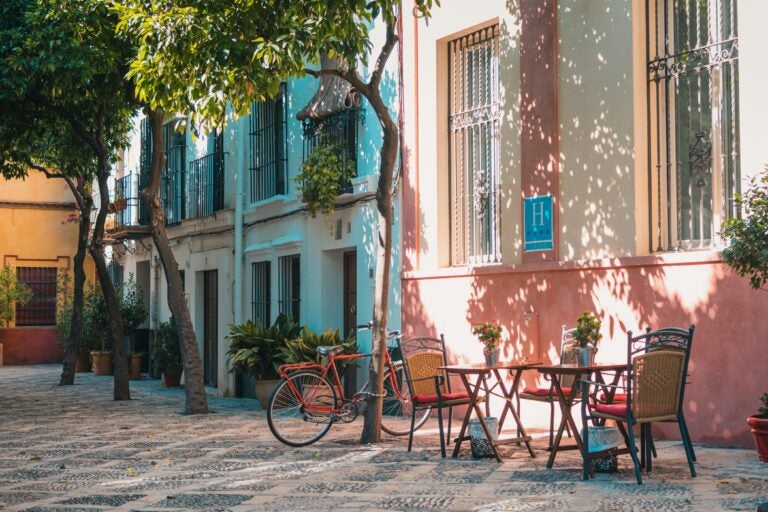 A sunny side street in Seville, Spain, with outdoor cafe tables, trees growing out of the pavement, and a bike resting against one of the trees.
