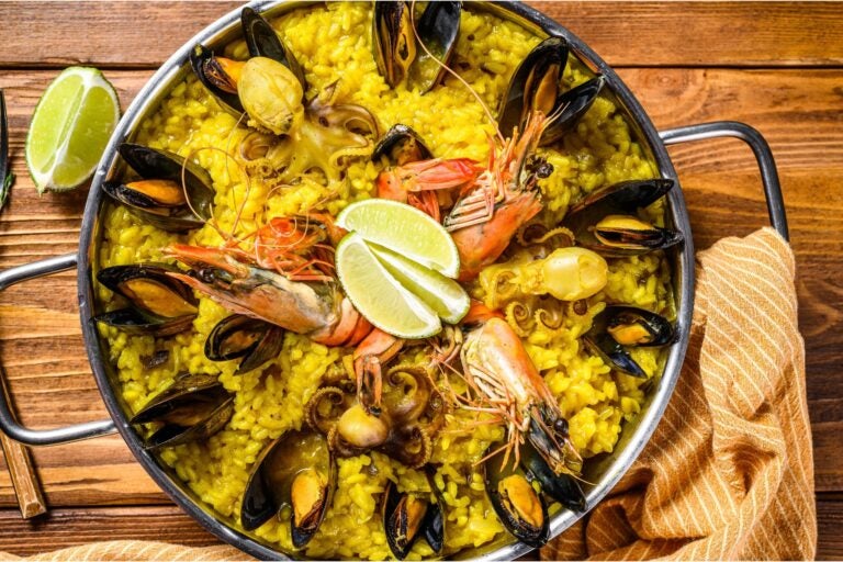 Up close and above shot of paella atop a wooden table, with an orange and white striped tea towel on the right handle of the metal dish.