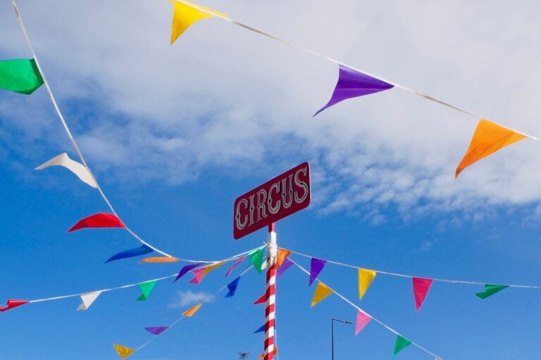 Upward outdoor shot of a red sign with "circus" written in white letters amid a blue sky with some cloud. Long white strings with small colorful flags drape off the sign's candy-stripe pole.