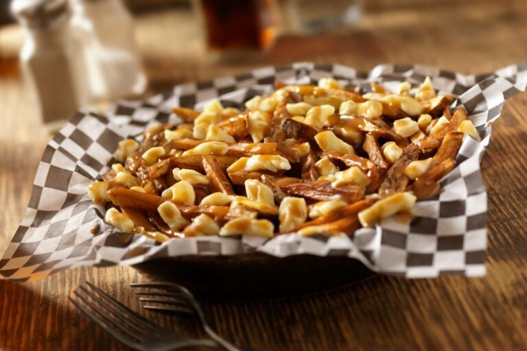 Closeup shot of the dish poutine. It sits atop a wooden table and in a bowl with black and white squared paper.