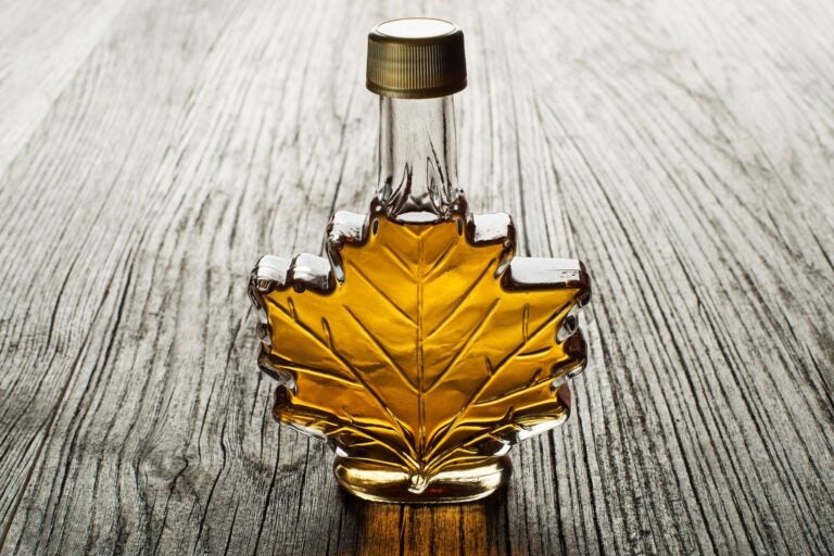 Glass bottle of maple syrup in the shape of a maple leaf atop a wooden table.
