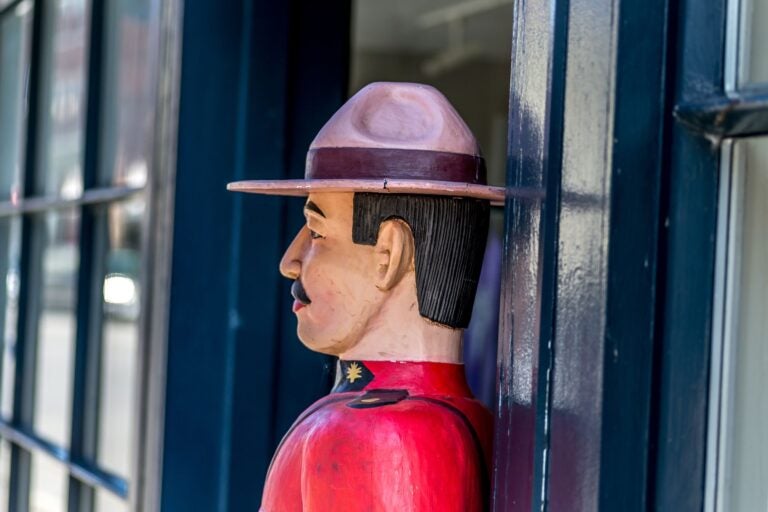 Side shot of a wooden carved Royal Canadian Mounted Police officer. The carved man is up against a blue wooden doorway.