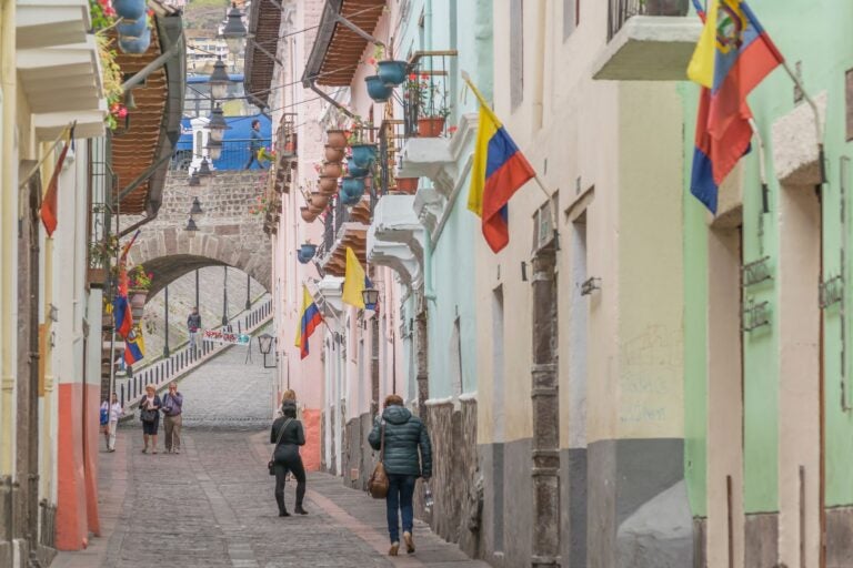 Cloudy day shot of the city of Quito, Ecuador, with people walking and standing. The Ecuadorian flag hangs off the side of buildings. 