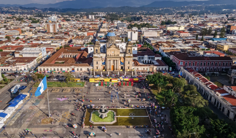 Aerial day shot of Guatemala City that is dotted with people and the Guatemalan flag to the left of the shot.