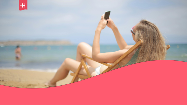 A young woman is lounging on a sunbed at the beach, holding her smartphone up to take a vacation selfie.