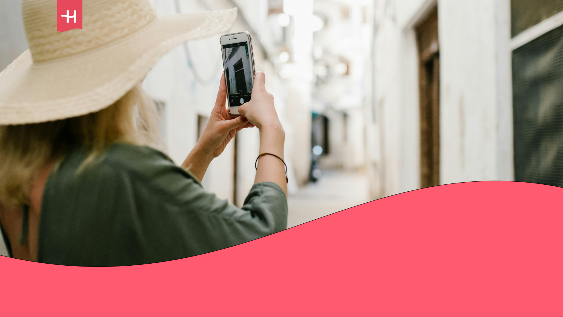 A tourist wearing a green dress and a wide-brimmed sun hat is taking a picture with her smartphone while exploring picturesque, narrow streets at her destination.