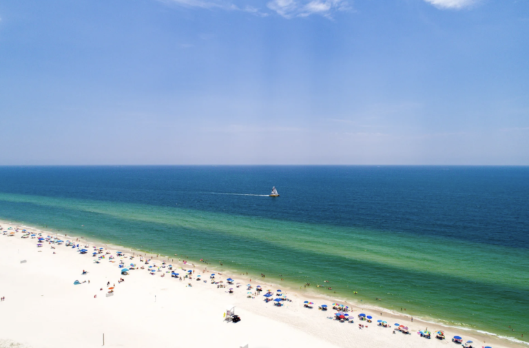 Aerial shot of Gulf State Park, Alabama on a sunny day with people lining the shoreline and a boat out in the sea.
