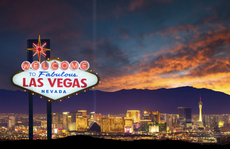 Evening shot of the Las Vegas city skyline and the "Welcome to Las Vegas Nevada" neon sign centre left of the shot.