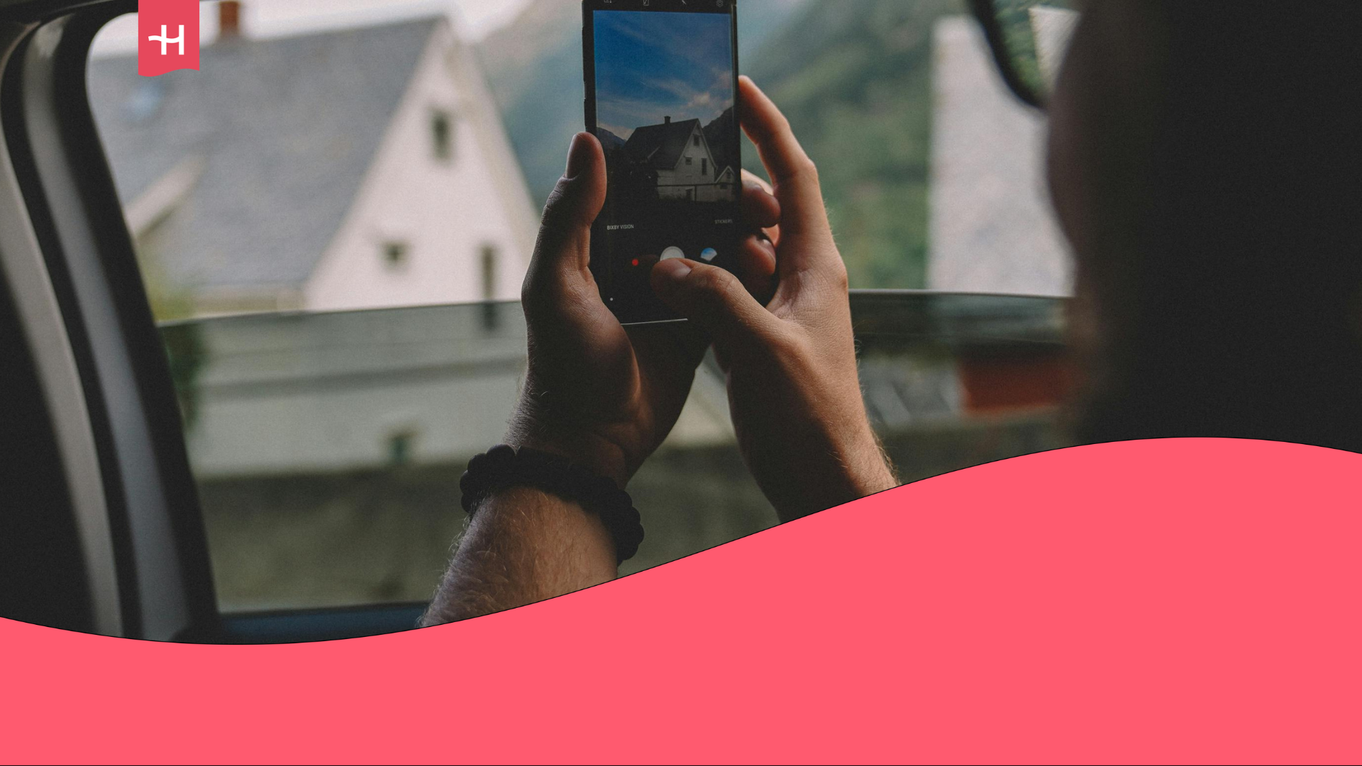 A traveler is sitting in the backseat of a cab, capturing a photo of picturesque homes nestled between mountains through the window using a smartphone.