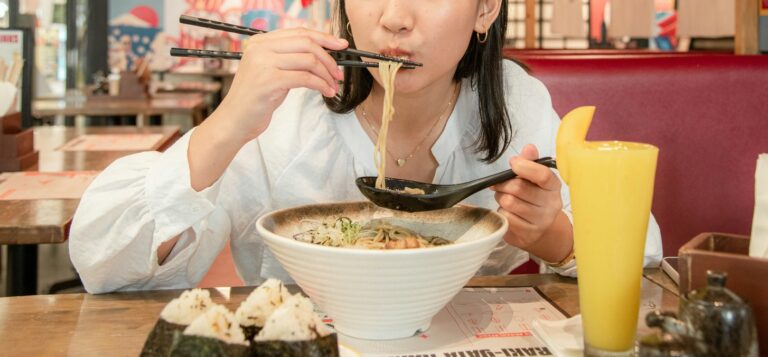 woman eating noodles with a chop stick and soup spoon from a large bowl of noodles 