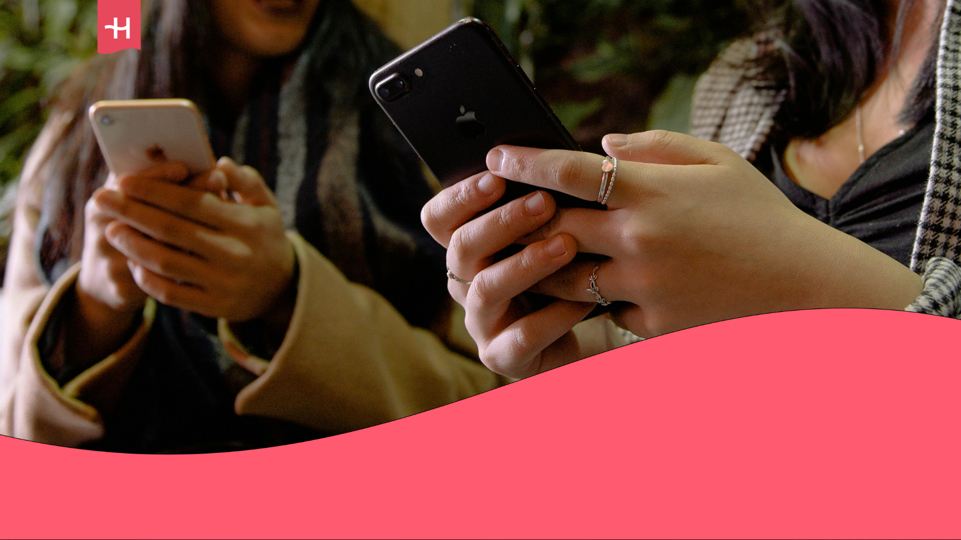 Two women are sitting side by side, each holding an iPhone. One woman is showing her phone screen to the other, who is laughing in response.