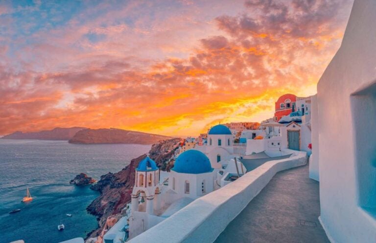 the iconic white-washed blue buildings in santorini with water below against a golden sunset 