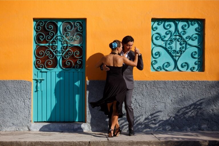 A couple dancing tango in front of a colorful yellow building with blue door and window shutters.