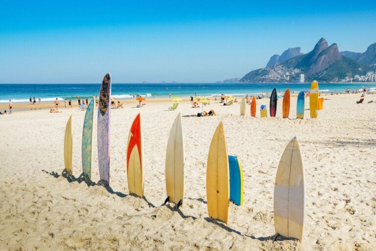People bathing and walking amidst colorful surf boards wedged in the sands of Ipanema beach, with mountains in the background.