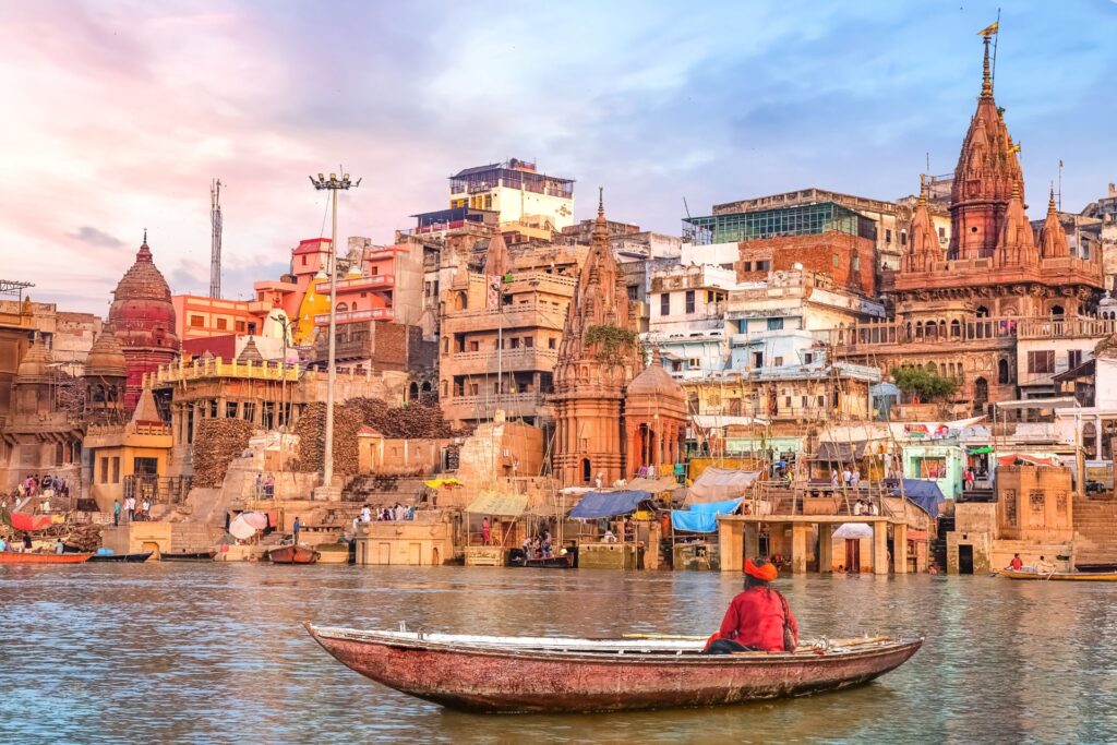 Man sitting in a small boat along a river facing the buildings of Jaipur, India.