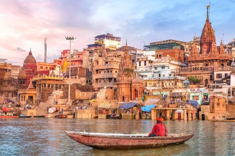 Man sitting in a small boat along a river facing the buildings of Jaipur, India.