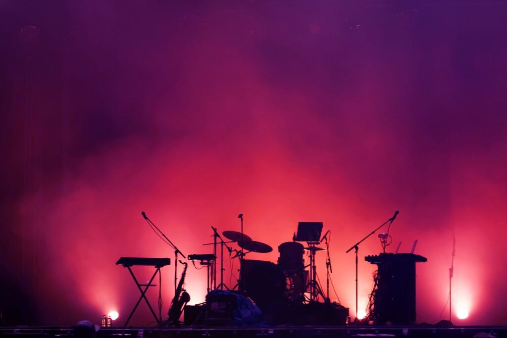 Shadow of a drum kit and keyboard in front of a pink and purple lit smoky lighting.