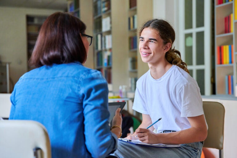 A high school student sitting down and talking to his guidance counselor while taking notes in a notebook.