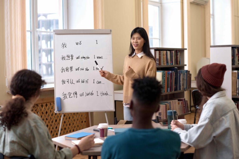A group of students in a Chinese language class watching as their language teacher points at some writing on a whiteboard.