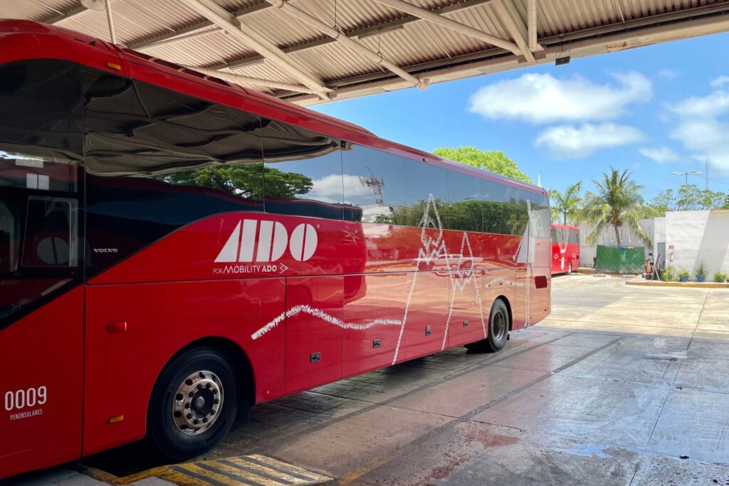 Side view of a red ADO bus stationed at an undercover bus terminal during the day.