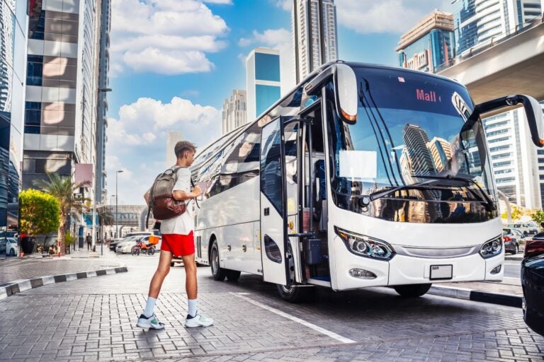 White bus stationed out front of a set of buildings. Man with red shorts, white t-shirt and brown backpack walking towards the bus.